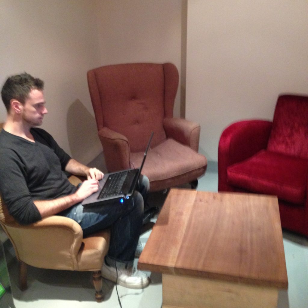 Assorted armchairs and a man working on a laptop perched on his knees