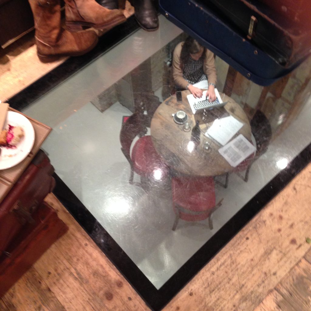 A small table for four occupied by one person on a laptop, viewed from above through a window in the floor